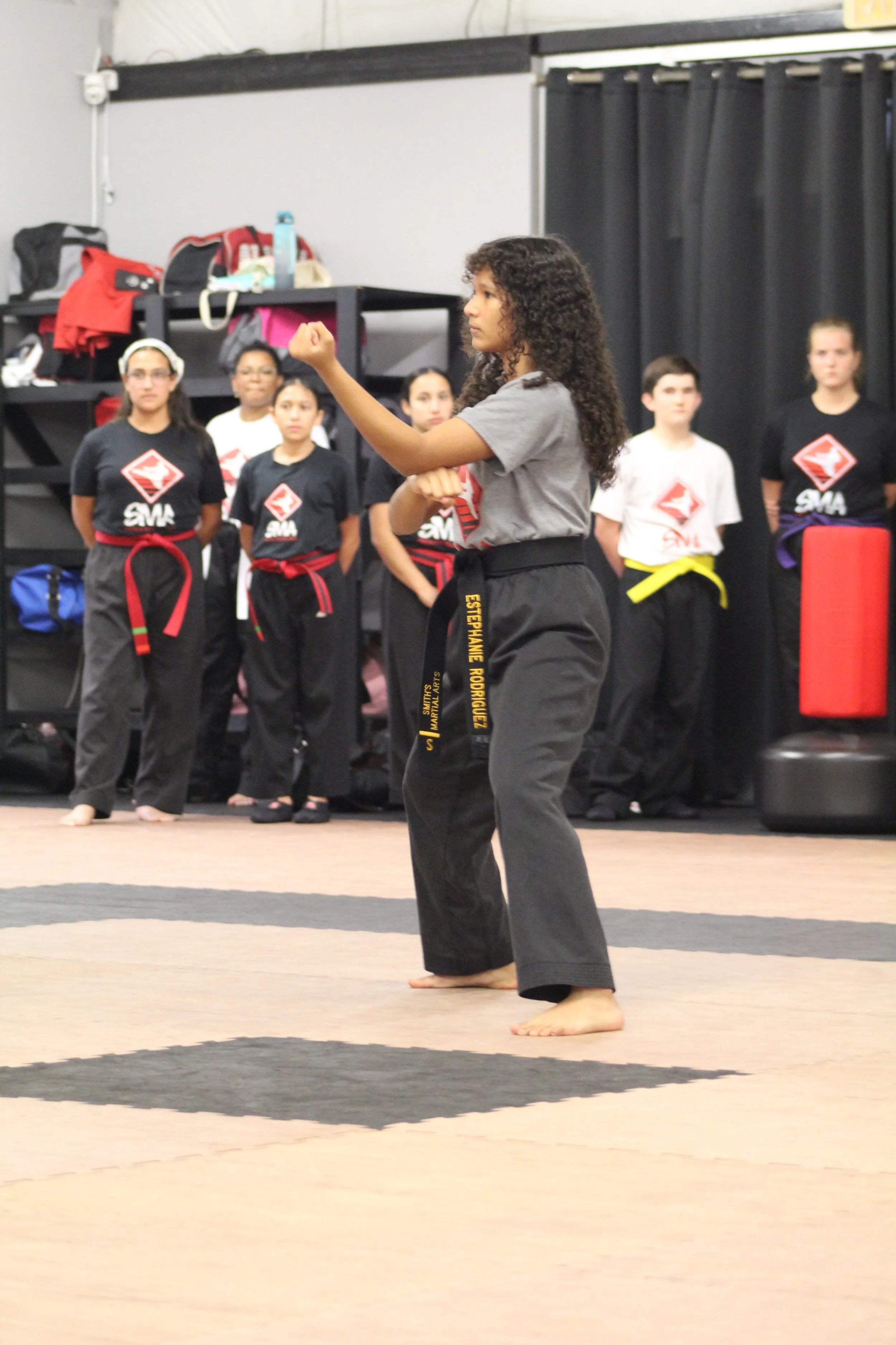A group of martial arts students with black belts watching a girl demonstrate a karate move in a dojo.