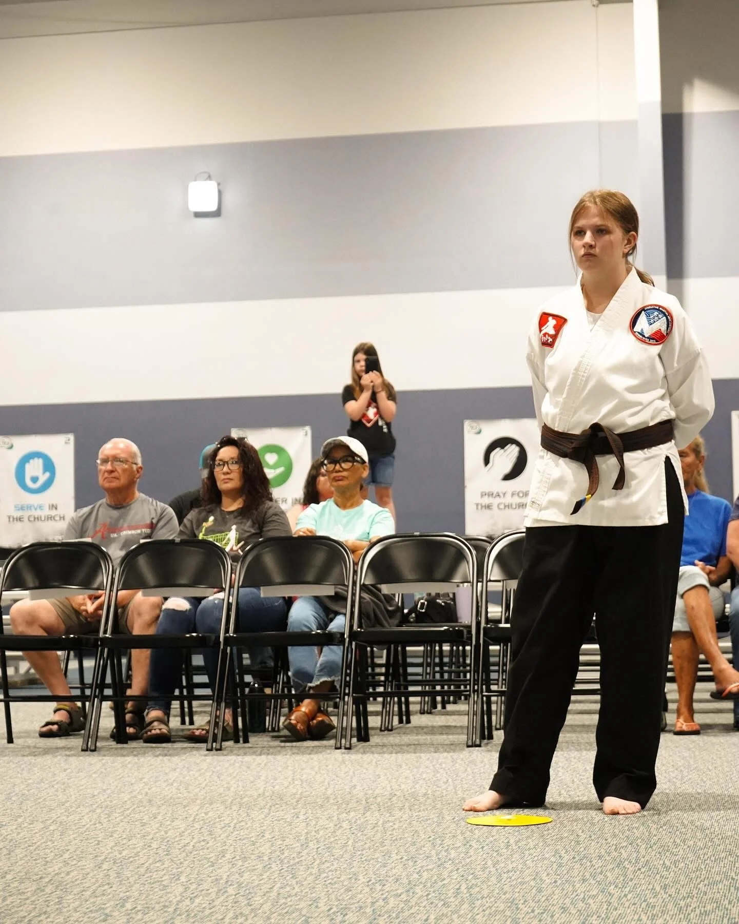 A young woman wearing a white martial arts uniform with patches and a brown belt standing barefoot in front of seated people at a martial arts event or class. Behind her, several people are seated. One girl is standing on a chair taking a photo. The 