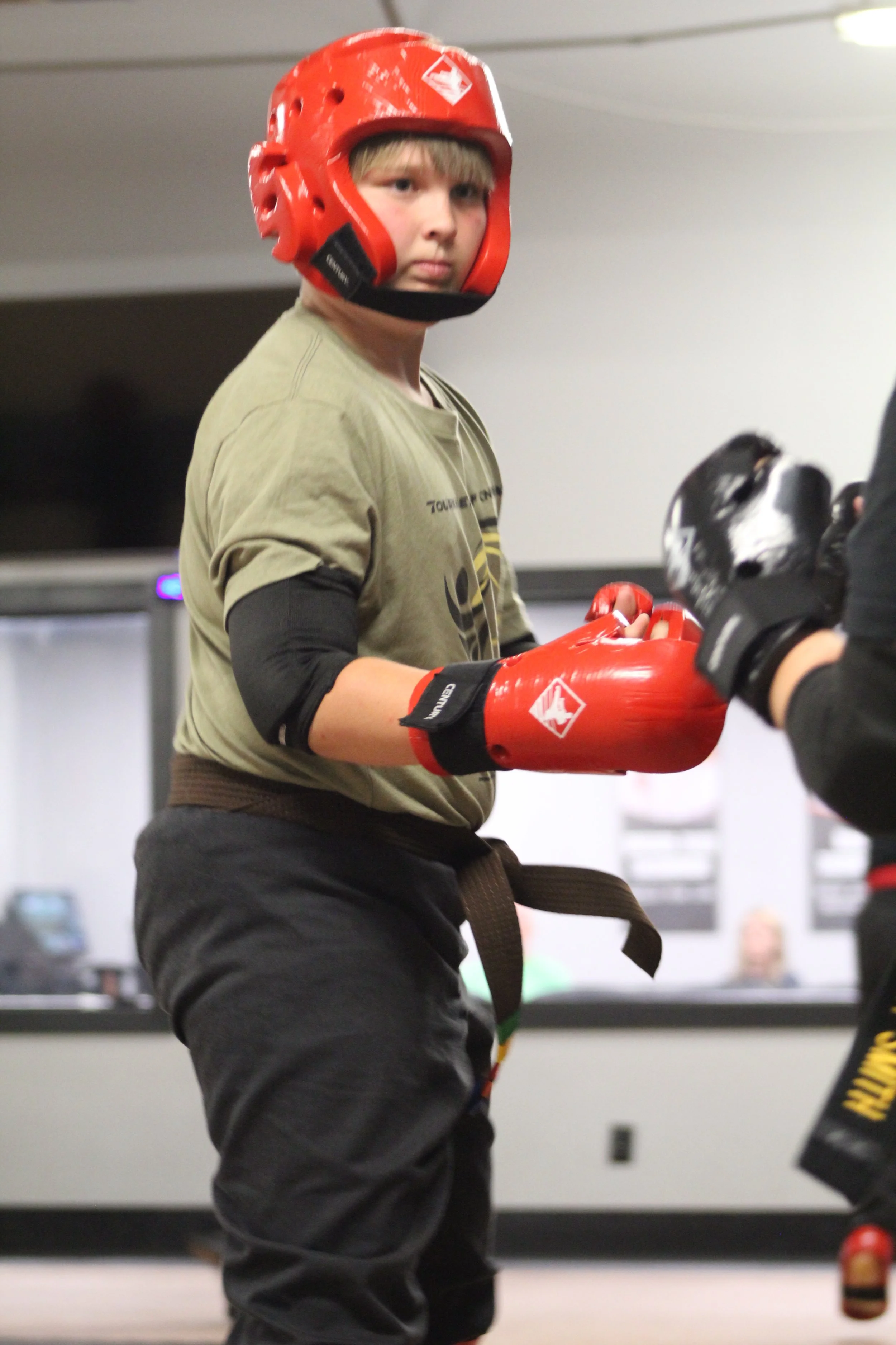 A young boy in martial arts gear wearing a red helmet and red gloves, holding a practice pad while standing in a martial arts training room.