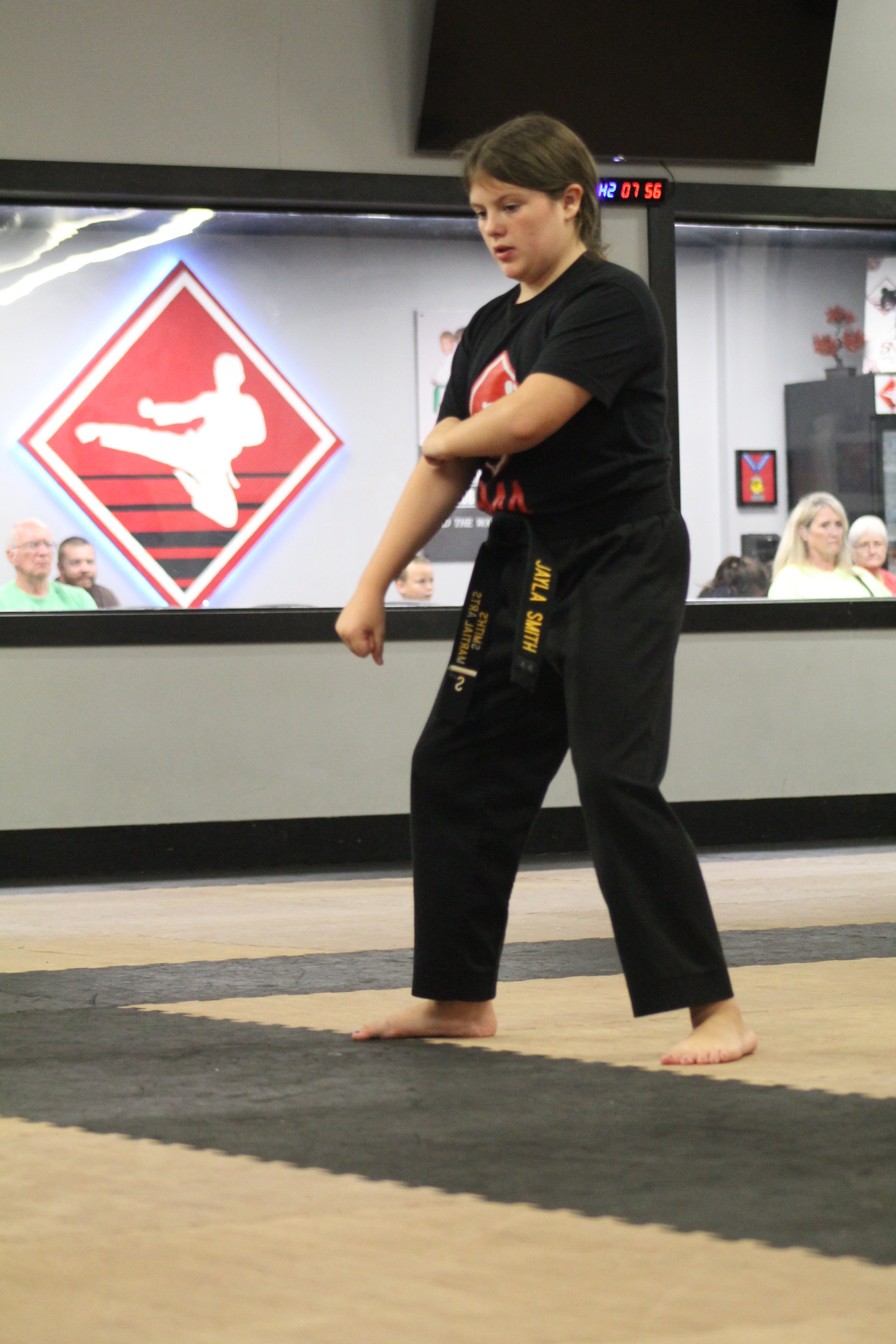 A young girl in a black martial arts uniform practicing martial arts on a mat in a dojo, with spectators watching behind a window.