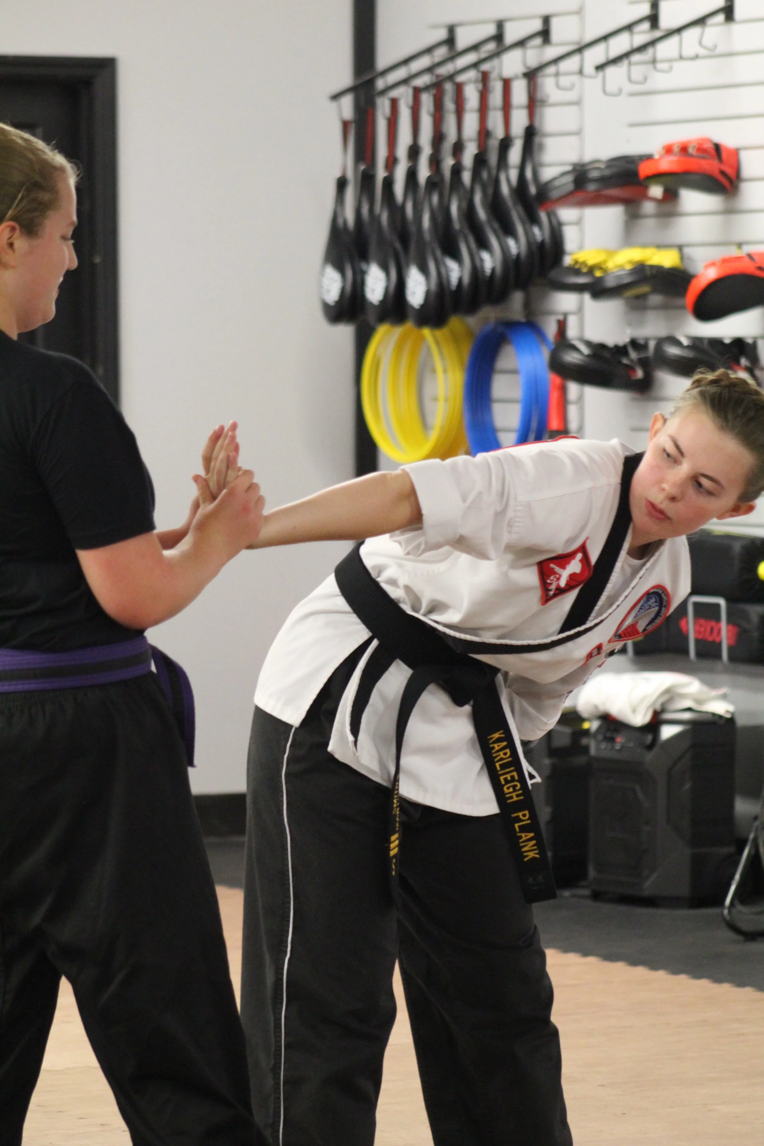 Two women practicing taekwondo in a dojo, with one woman holding the other back with a martial arts move.