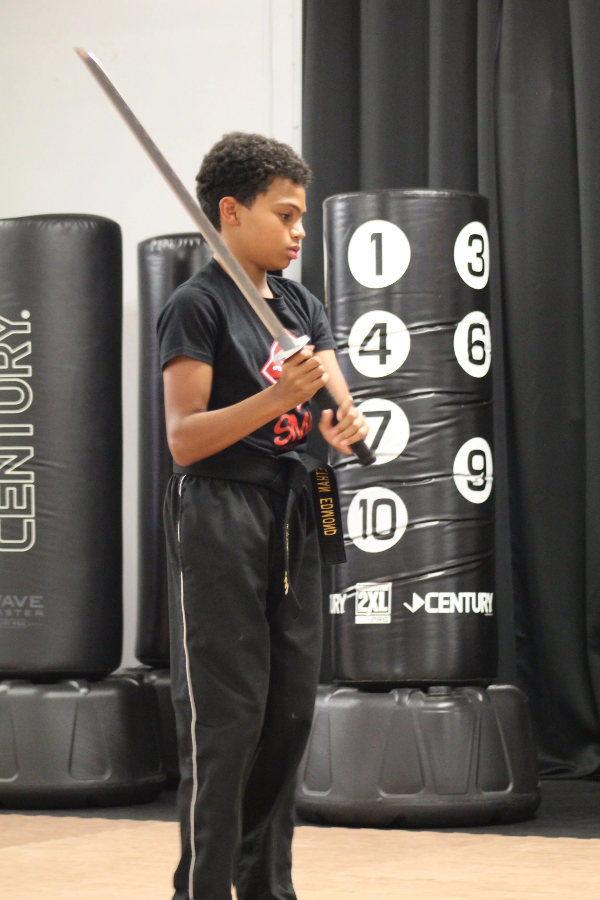 A young boy wearing a black martial arts uniform and a black belt holding a sword in a martial arts or boxing gym. Behind him are stacked punching bags with numbers.
