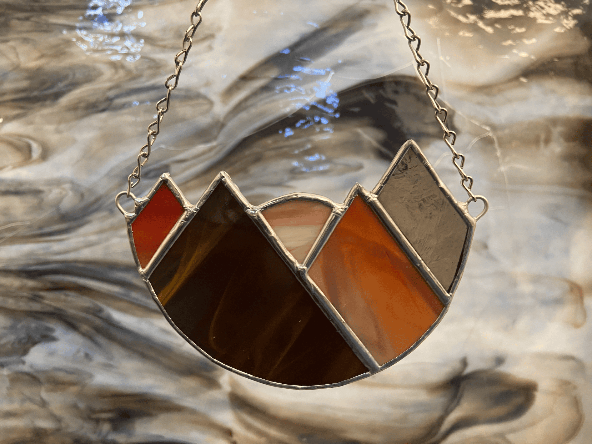 A warm-toned stained glass mountain with a sun setting hangs on a chain in front of a background of brown and white swirled glass