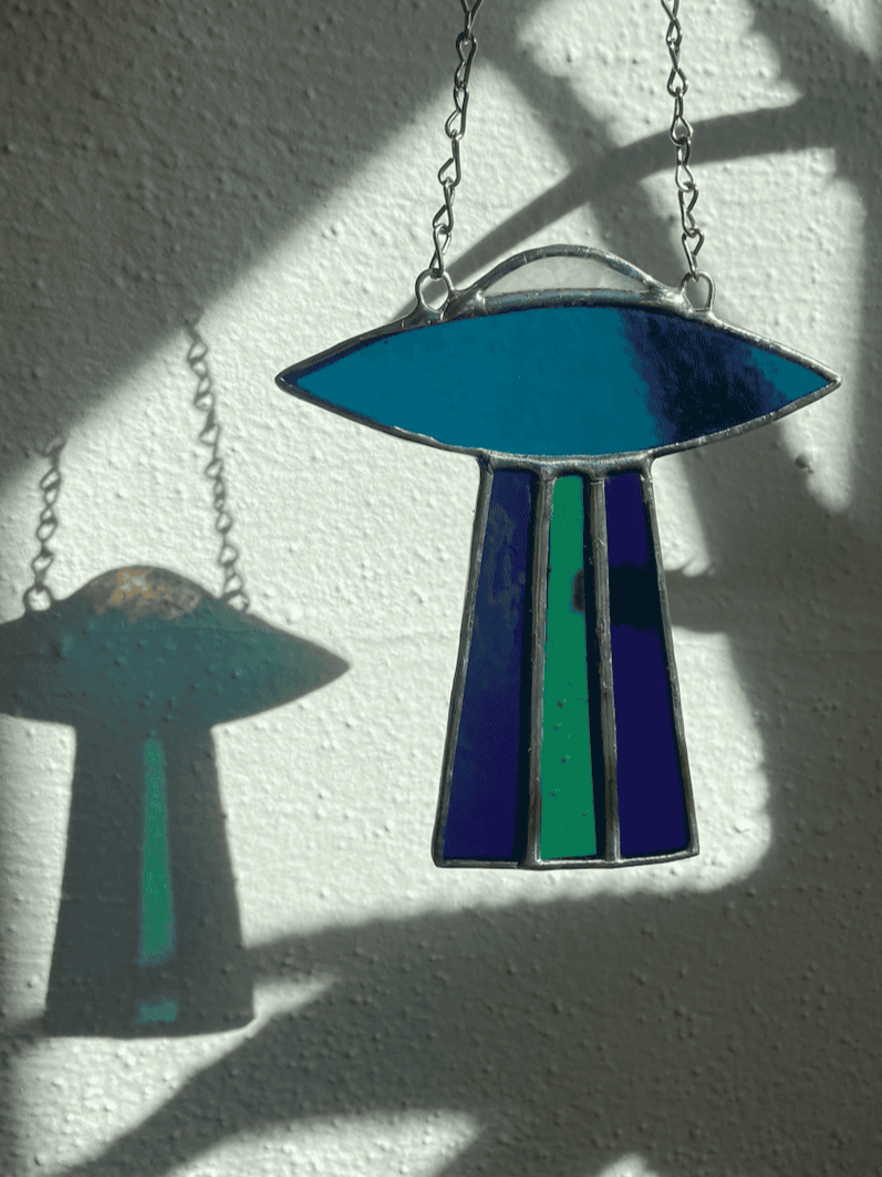 A turquoise and blue UFO stained glass suncatcher hangs from a chain as light passes through and creates a colorful shadow on the wall
