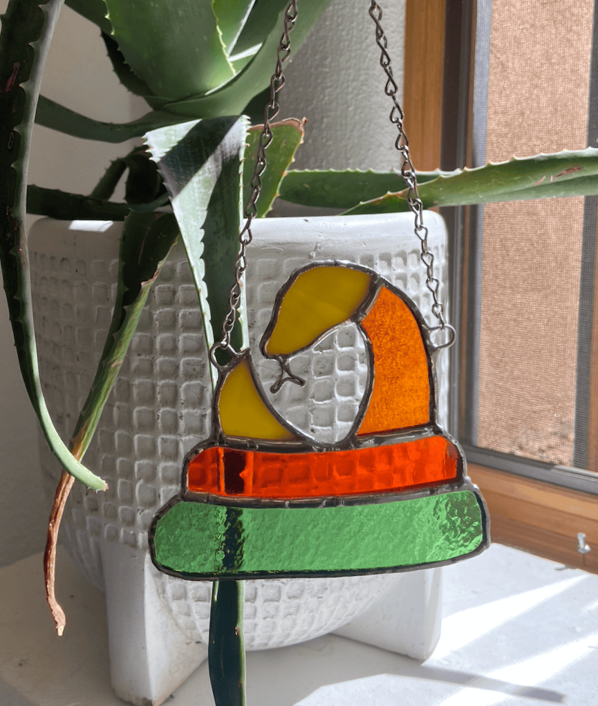 A brightly colored stained glass snake hangs on a chain in front of a potted aloe plant as sunlight from a window illuminates the yellow, orange, and green glass pieces