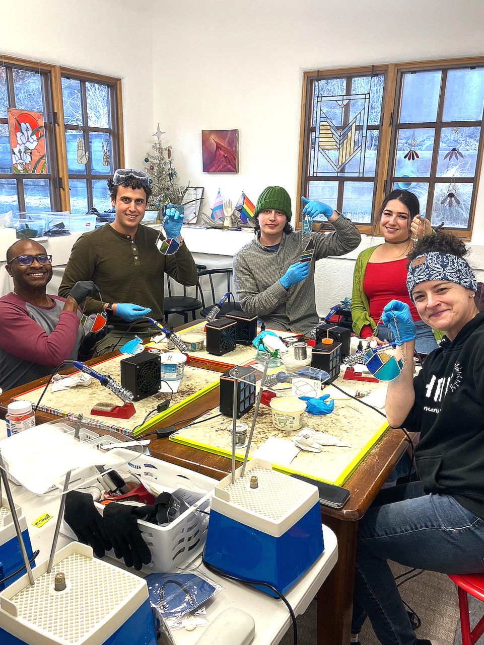 Five smiling friends show off their stained glass suncatcher projects while sitting at their workstations in a studio classroom with stained glass panels hanging in the background