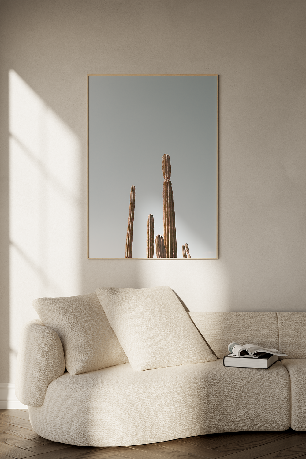 A beige textured sofa with a pillow and a book on top, in a living room, with a framed wall art of cacti and sunlight casting shadows on the wall.