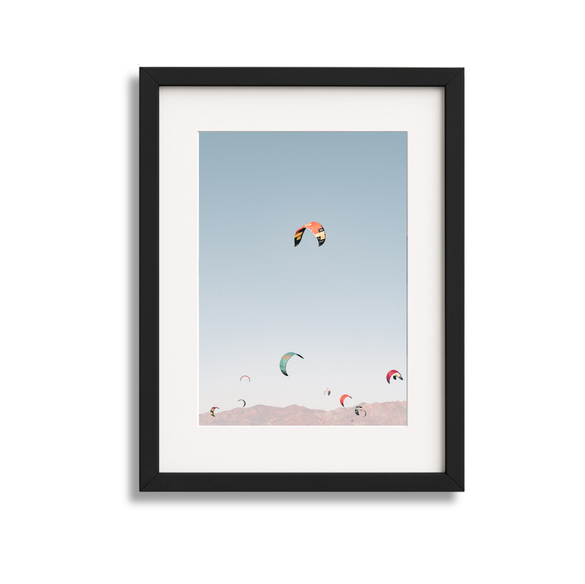 Framed photo of multiple colorful kite surfers flying kites over mountains on a clear sky day.