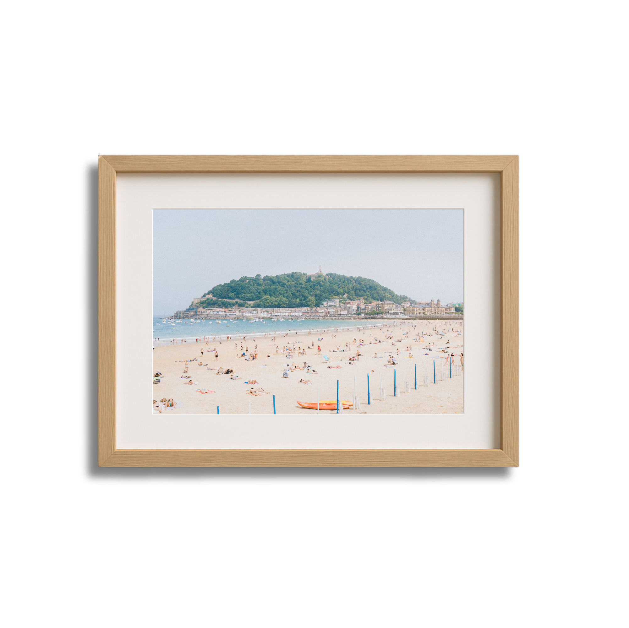 Framed photograph of a sandy beach with people sunbathing and swimming, a hill with greenery and buildings in the background, and a clear sky.