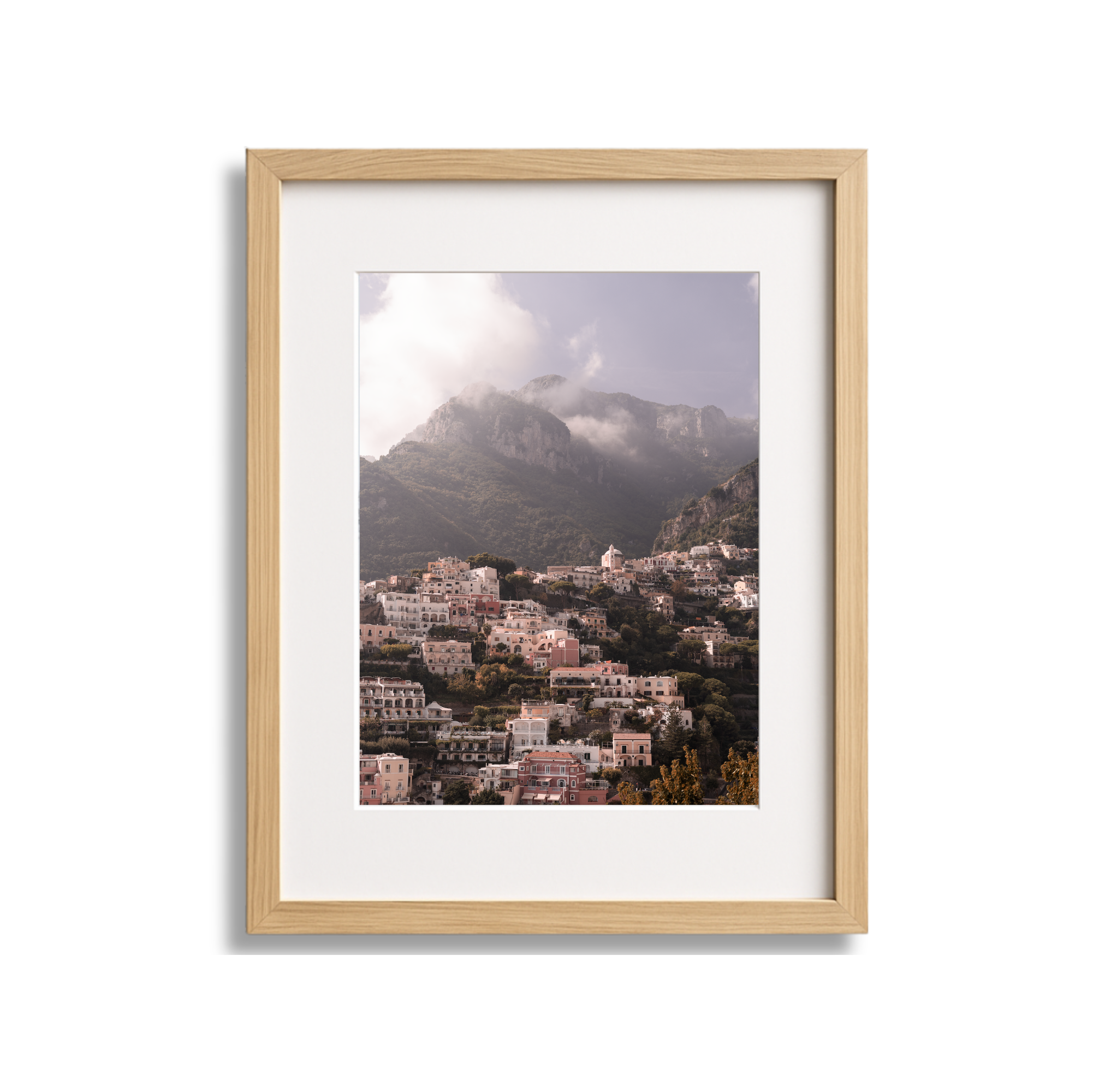 Framed landscape photograph of a hillside town with pastel-colored buildings and lush greenery, mountain range with cloudy sky in the background.