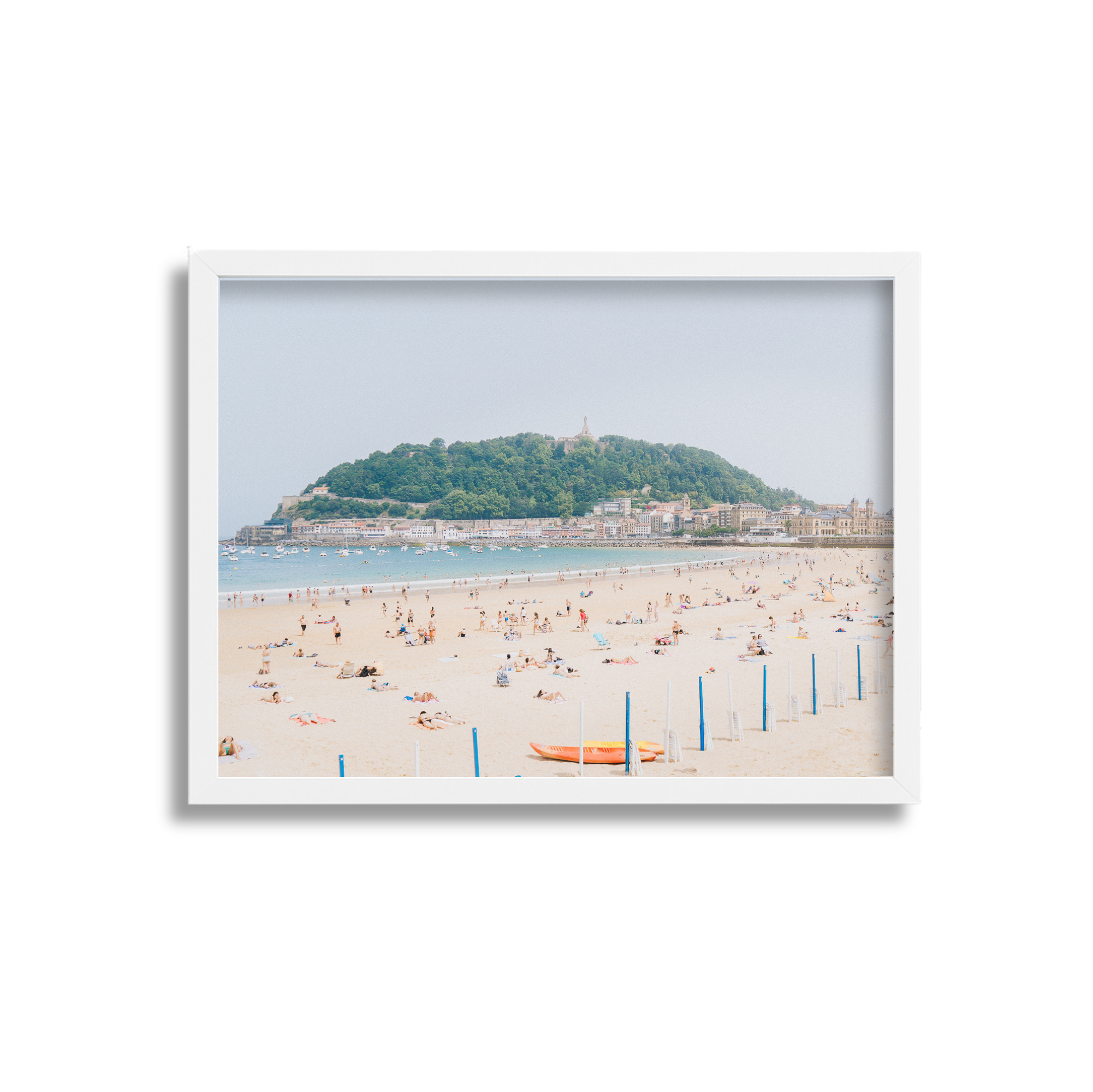 Beach with people sunbathing and swimming, green hill in the background, white buildings along the shoreline.