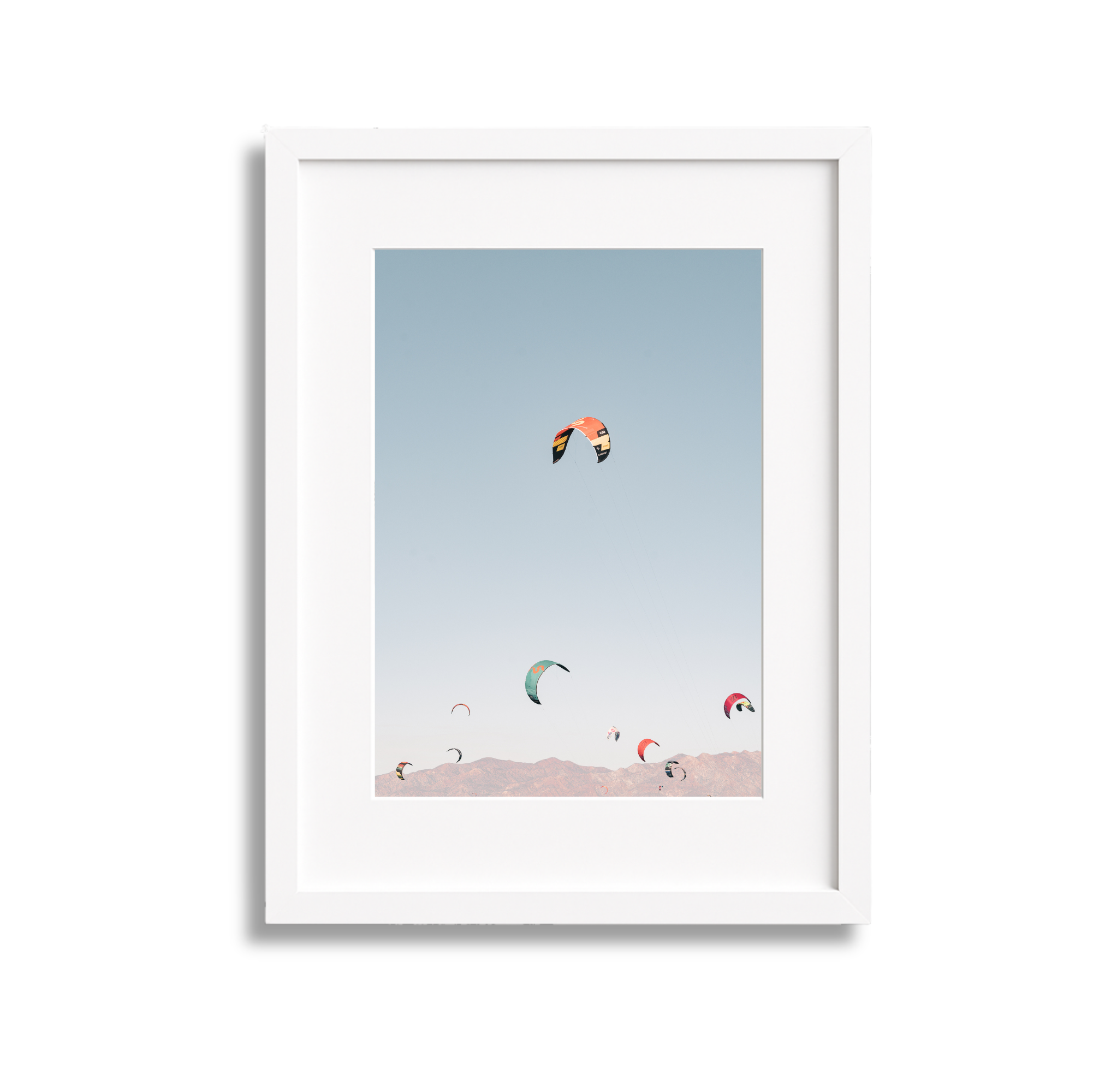 Framed photograph of multiple colorful kites flying in a clear sky over a mountainous landscape.