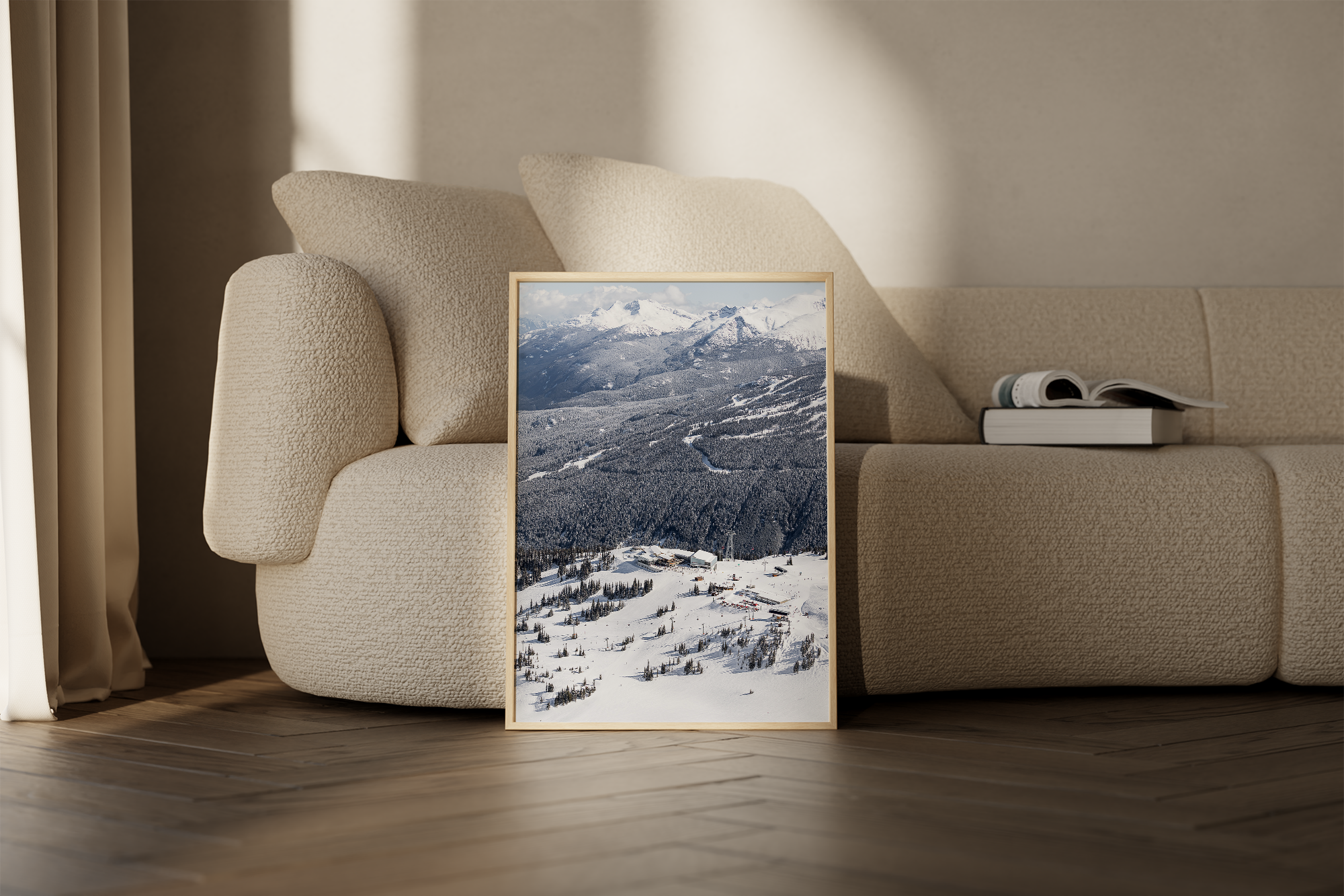 Framed picture of snow-covered mountains and a ski resort leaning against a beige sofa with pillows, with books on the sofa armrest.