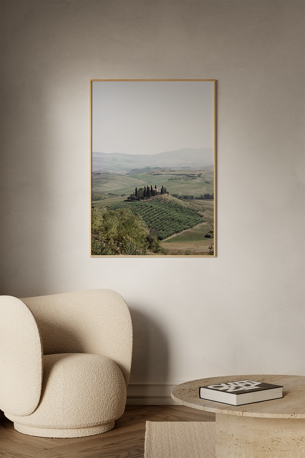 Living room with a beige textured armchair, round beige marble table with a black book, and a large framed landscape photograph of rolling hills and cypress trees on the wall.