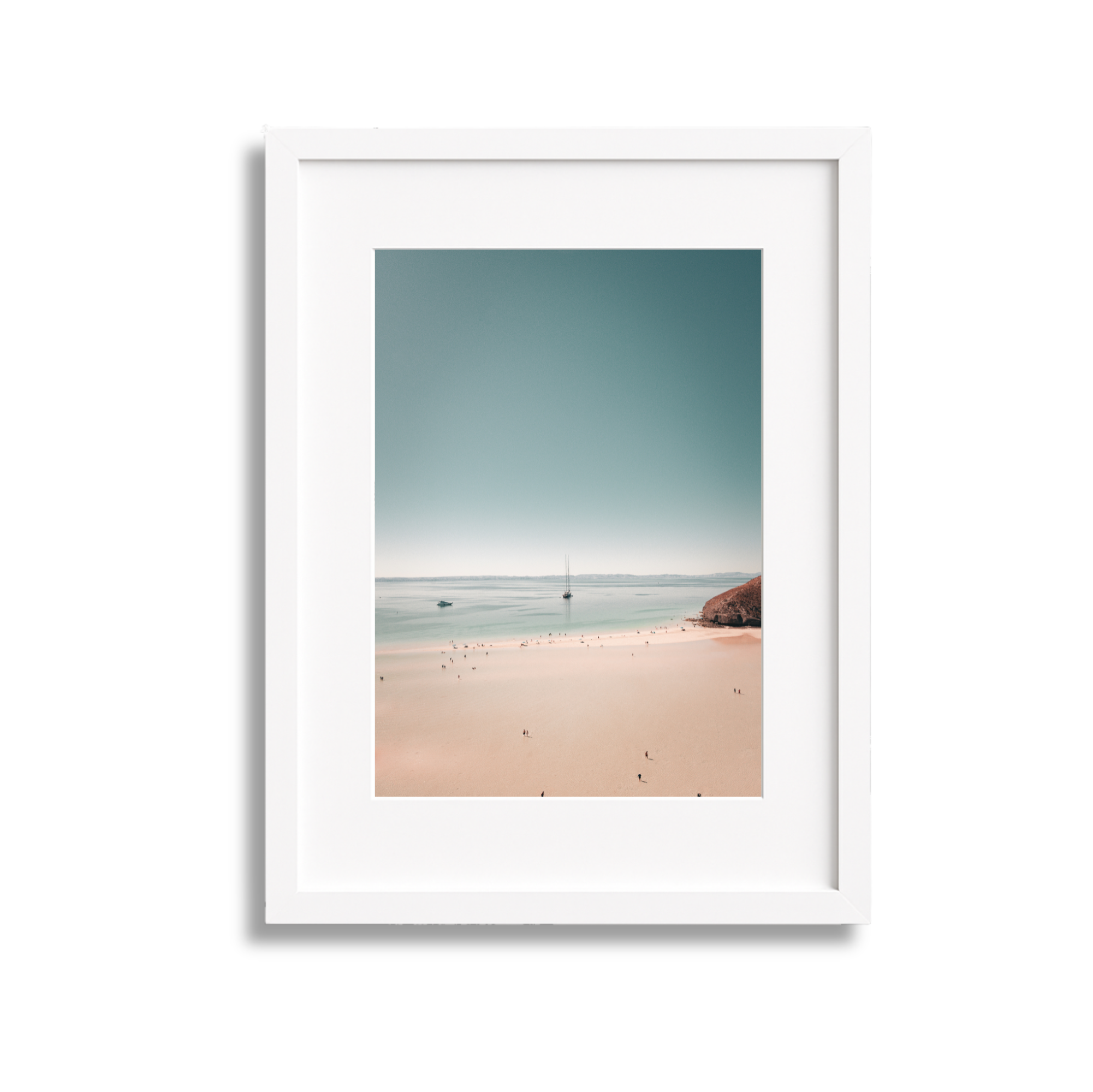 Photograph of a beach with sandy shoreline, small boats in the water, and a rocky hill on the right, framed in a white picture frame.