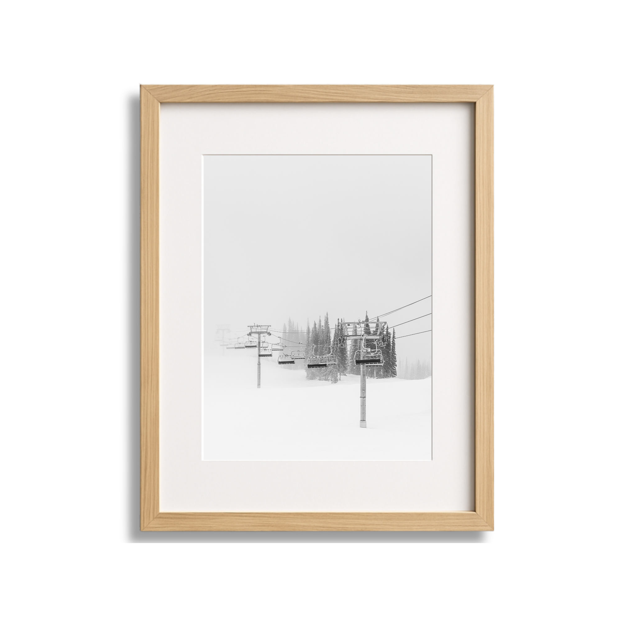 Black and white photograph of ski lift chairs on a snowy mountain with pine trees in the background, framed in a wooden frame.