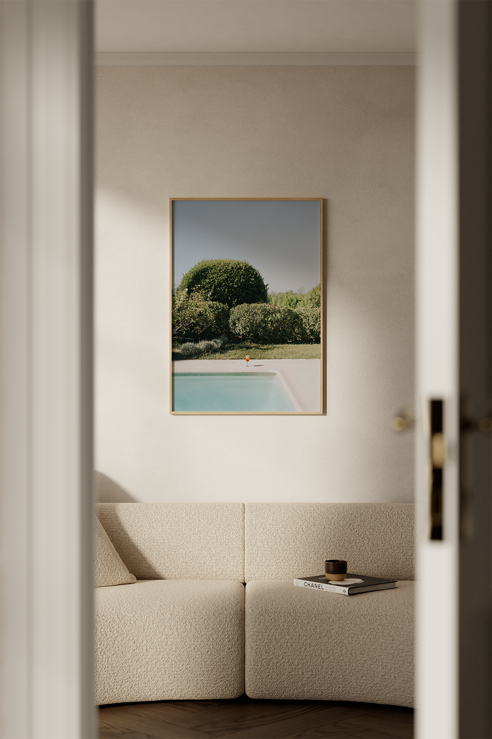 View through an open door showing a beige sofa with a pillow, a black bowl, and a white book titled 'Chanel' on it, with a framed landscape painting of trees, a swimming pool, and a clear blue sky hanging on the wall behind.