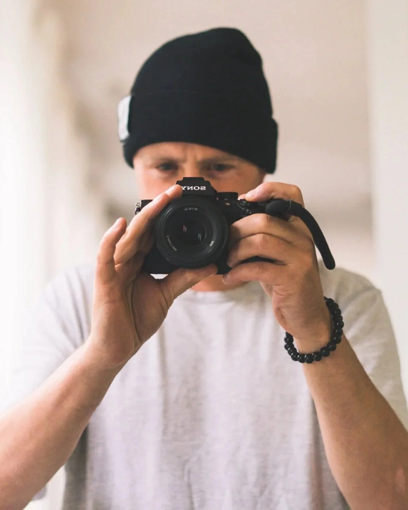 Person taking a photo with a Sony camera, wearing a black beanie and a grey t-shirt, in a mirror.