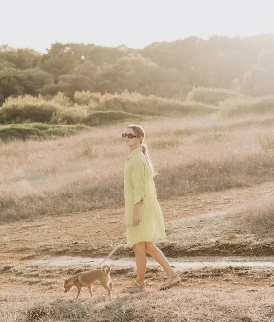 A woman in a yellow dress walking a small dog on a dirt path in a field with tall grass, with hills and trees in the background and sunlight in the sky.