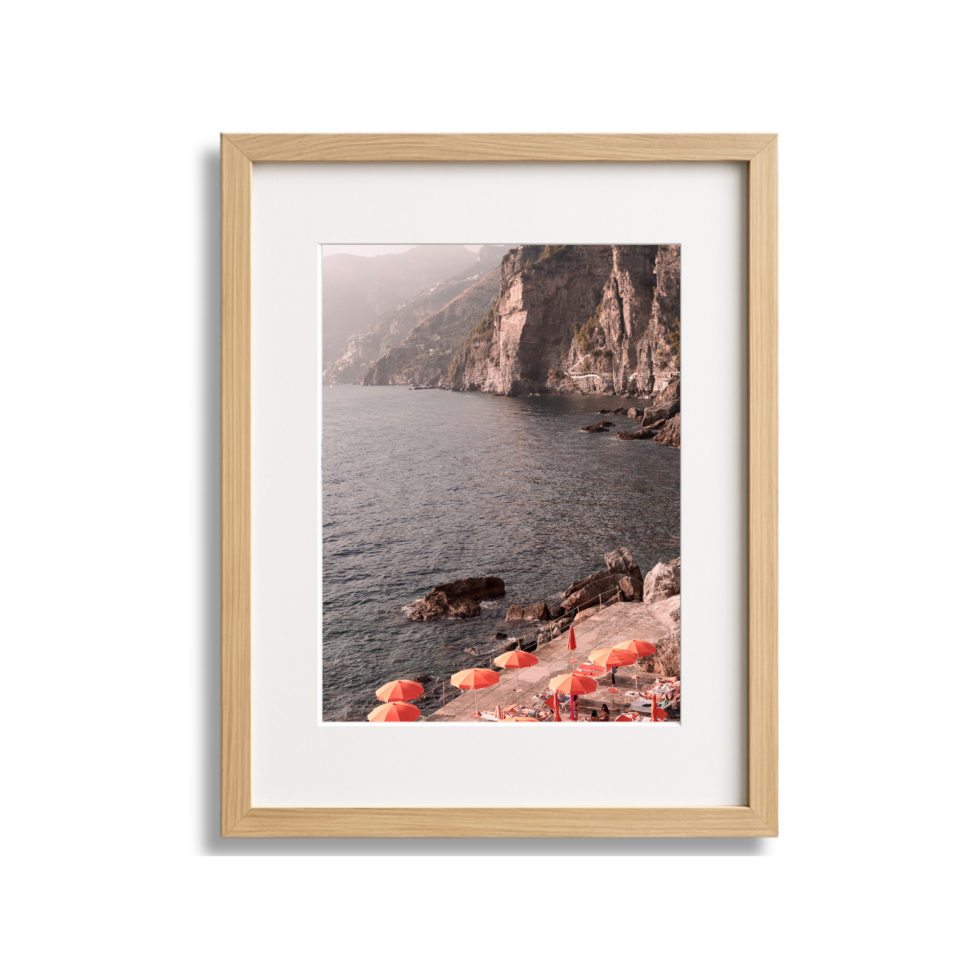 Framed photograph of rocky coastline with cliffs and water, featuring a beach with orange umbrellas and chairs.