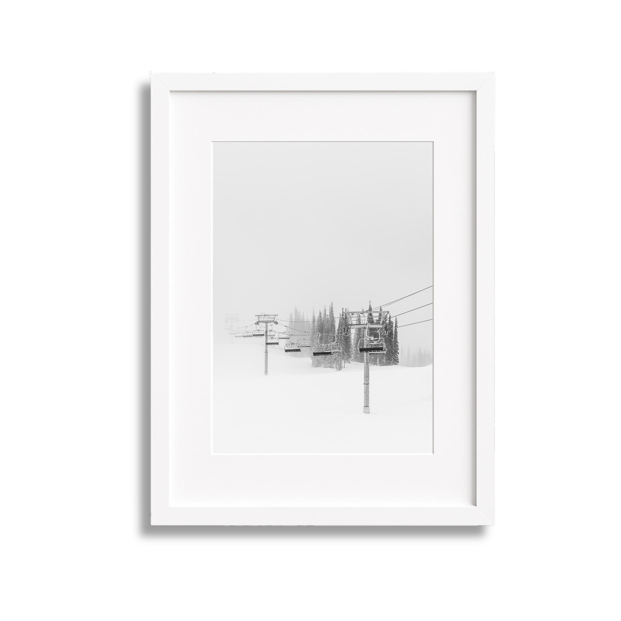 White picture frame containing a black and white photo of ski lift chairs and towers amid a snowy, foggy landscape with pine trees.