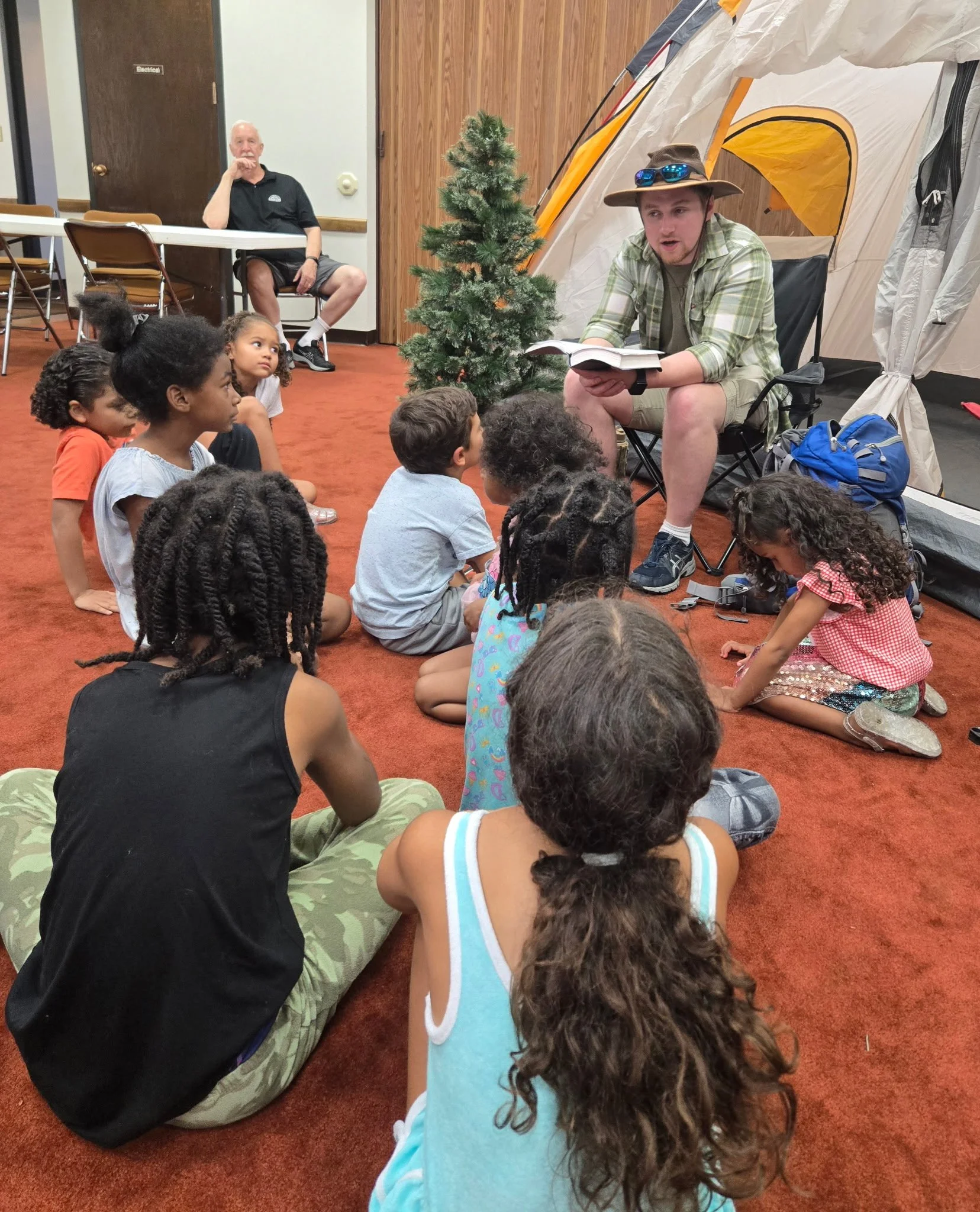 A man dressed as a ranger reading a book to a group of children sitting on the floor inside a tent at a camping or educational event, with a small artificial tree and a man seated at a table in the background.