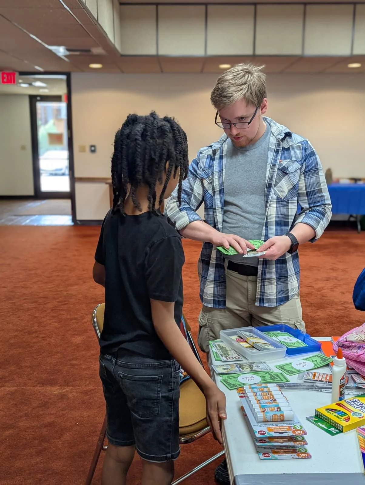 A man with glasses and a plaid shirt is handing money to a young boy with dreadlocks at a table, which has various school supplies, including cards, glue, and other stationery items.