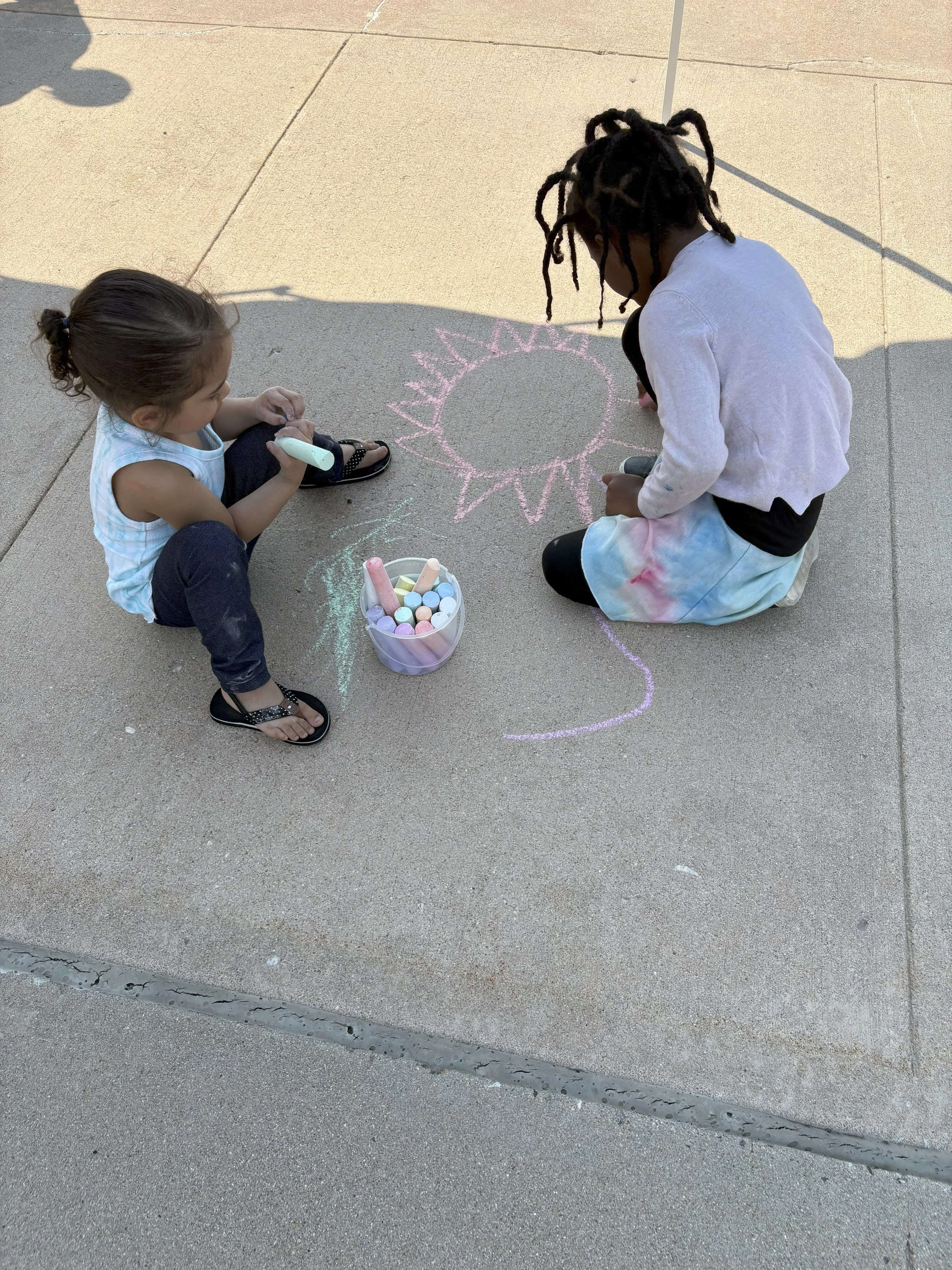 Two children drawing with chalk on pavement, with a bucket of chalk nearby, creating a sun.