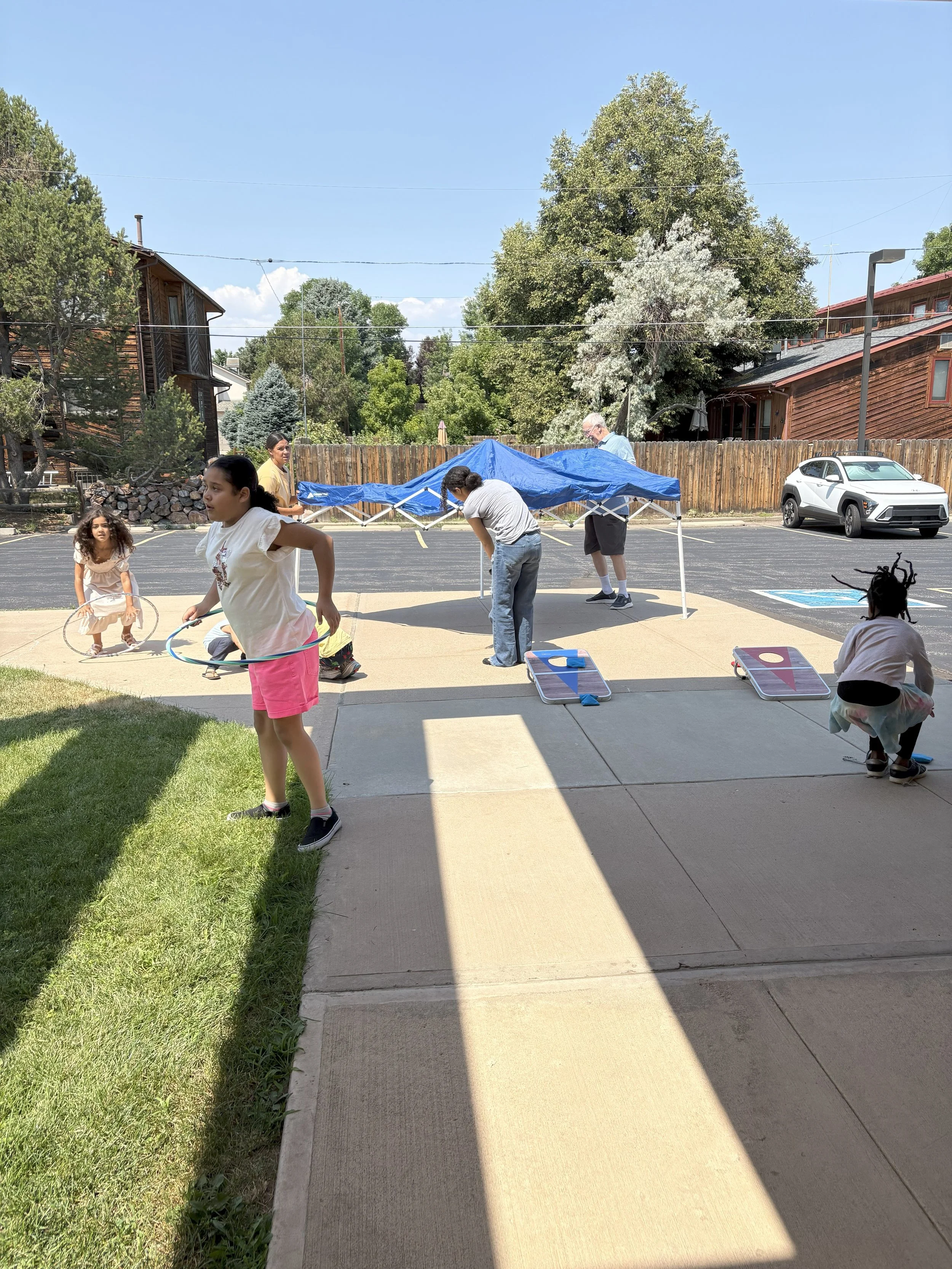 Children playing and setting up outdoor activities in a sunny yard with a tent, cornhole boards, and a parked car nearby.