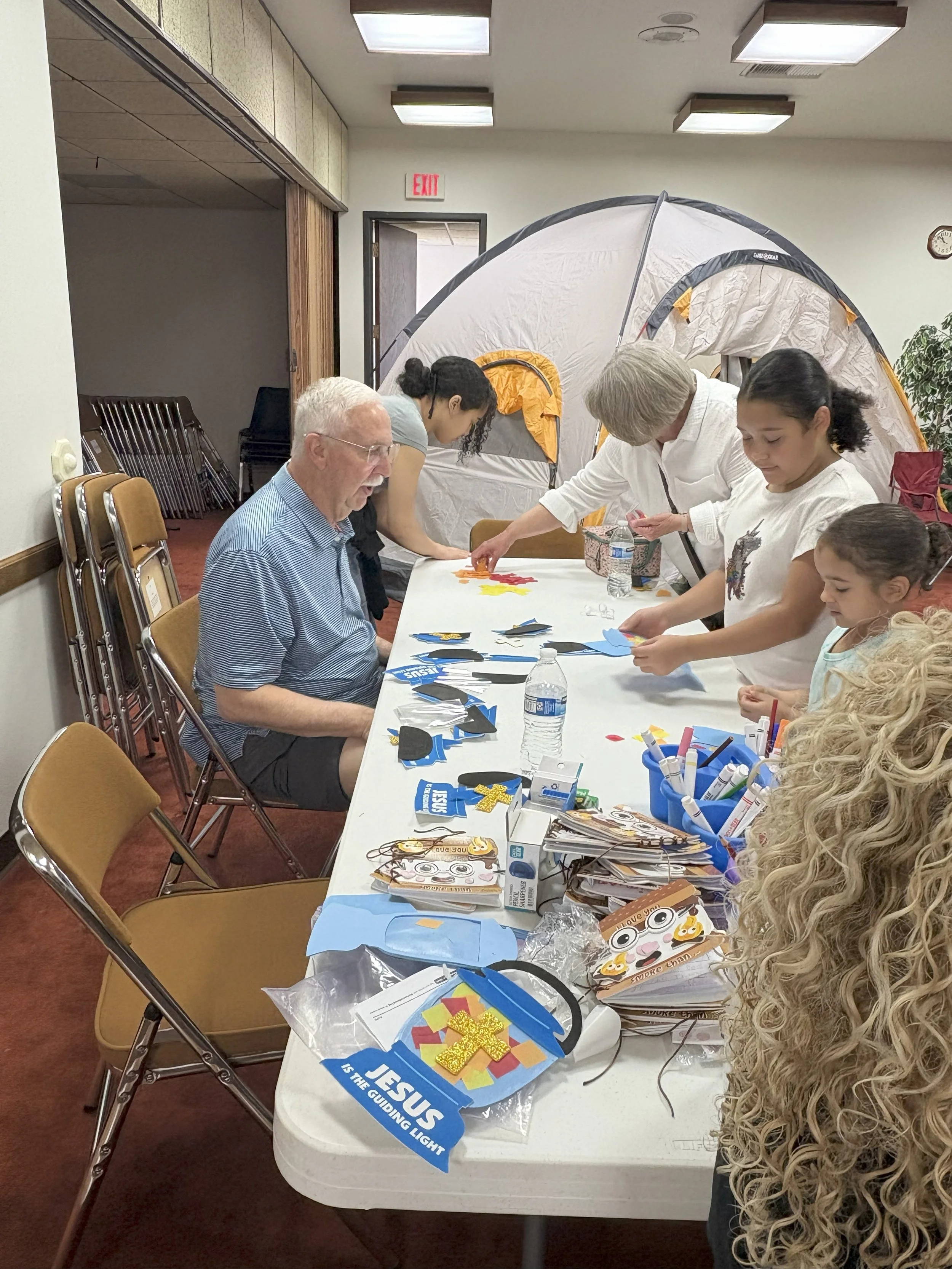 Group of people making craft projects at a long table. Supplies include foam, markers, and paper. A tent is set up in the background.