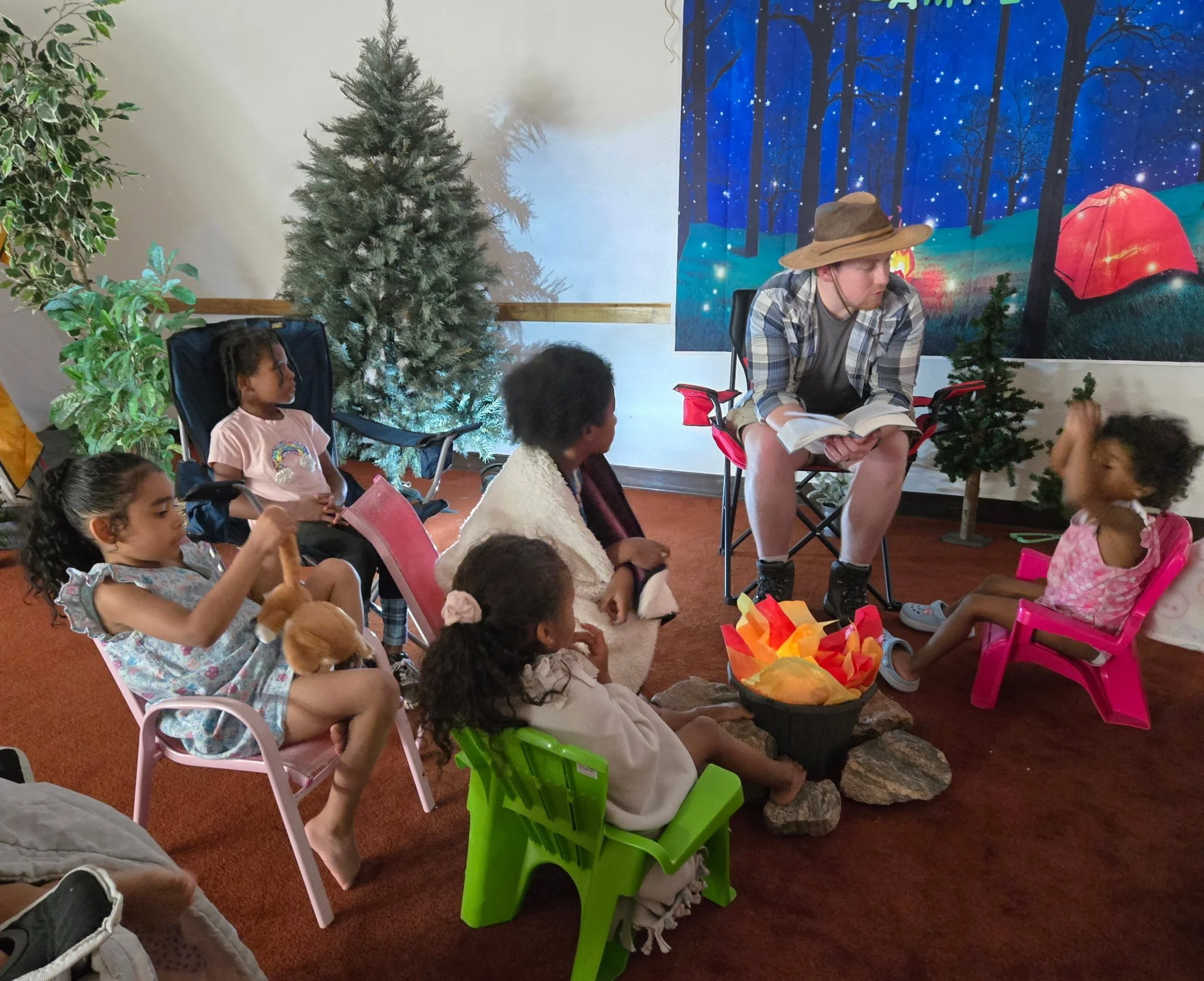 Youth storyteller in outdoor camping scene reading to children seated around a campfire set with artificial flames, in a room decorated with faux Christmas trees and a nature-themed backdrop.