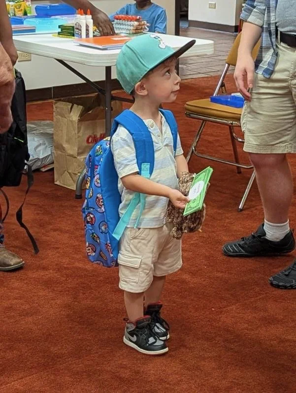 A young boy with a blue backpack, wearing a light gray cap, short-sleeved shirt, khaki shorts, and sneakers, standing indoors on a stained red carpet, holding a green pamphlet and a stuffed animal, surrounded by adults.