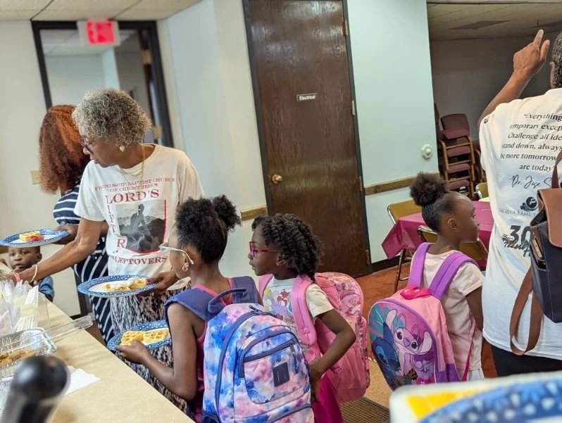 Children with backpacks waiting in line for food at a table in a community center or gathering space.