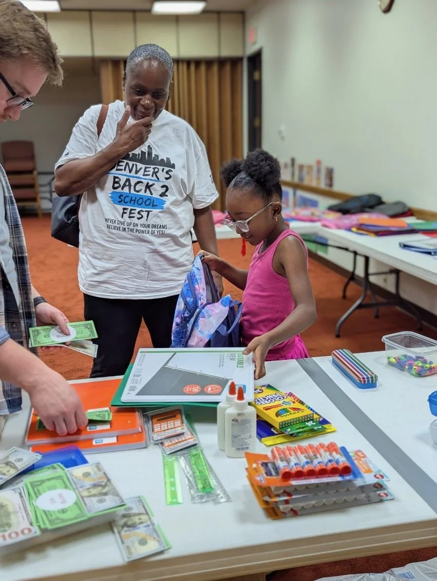 A young girl and two men at a table with school supplies and cash, likely in a school or community center. The girl is smiling with books and backpacks nearby.