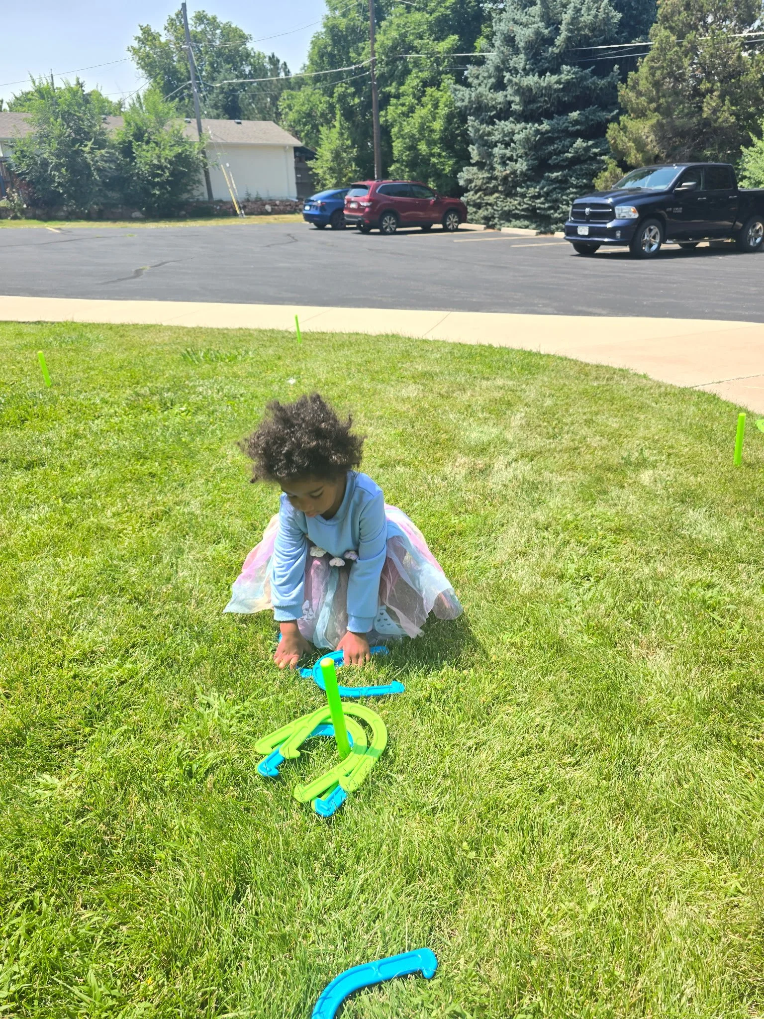 A young girl with curly hair playing with a plastic pool noodle and toy tools on a grassy lawn on a sunny day.