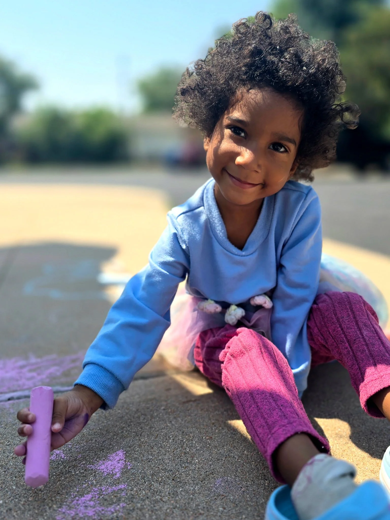 Young girl with curly hair drawing on pavement with pastel chalk, smiling at the camera.