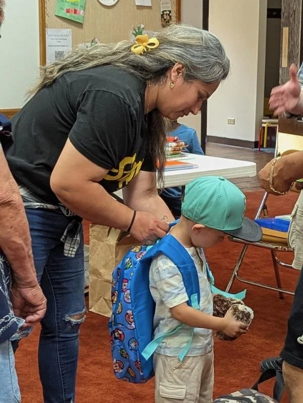 A woman assisting a young boy, who is wearing a cap and backpack, holding a chocolate cake in a room with books and chairs.