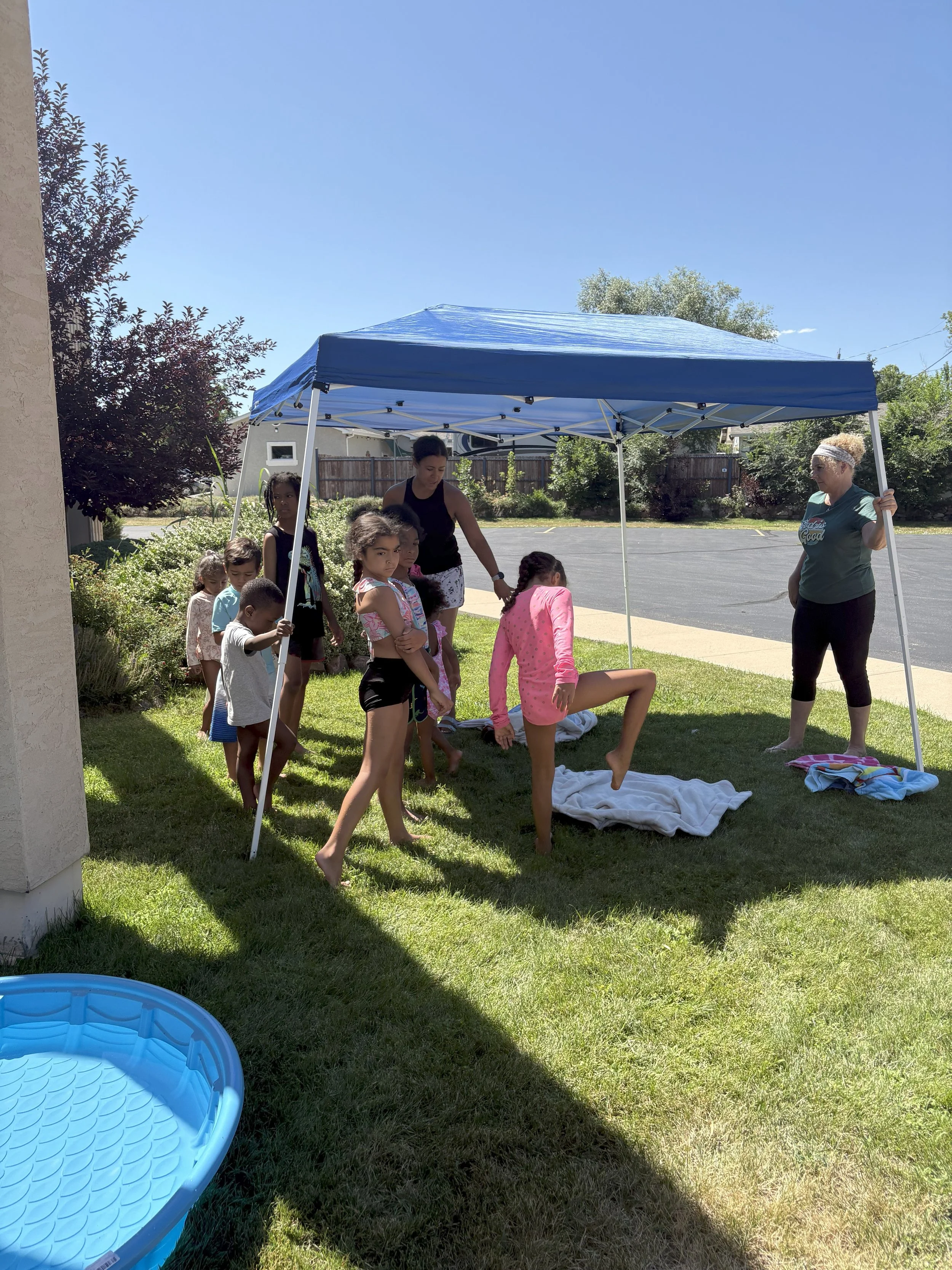 Children and adults gathered under a blue canopy outside on a sunny day, with children standing and one girl practicing a gymnastic move on a towel on the grass.