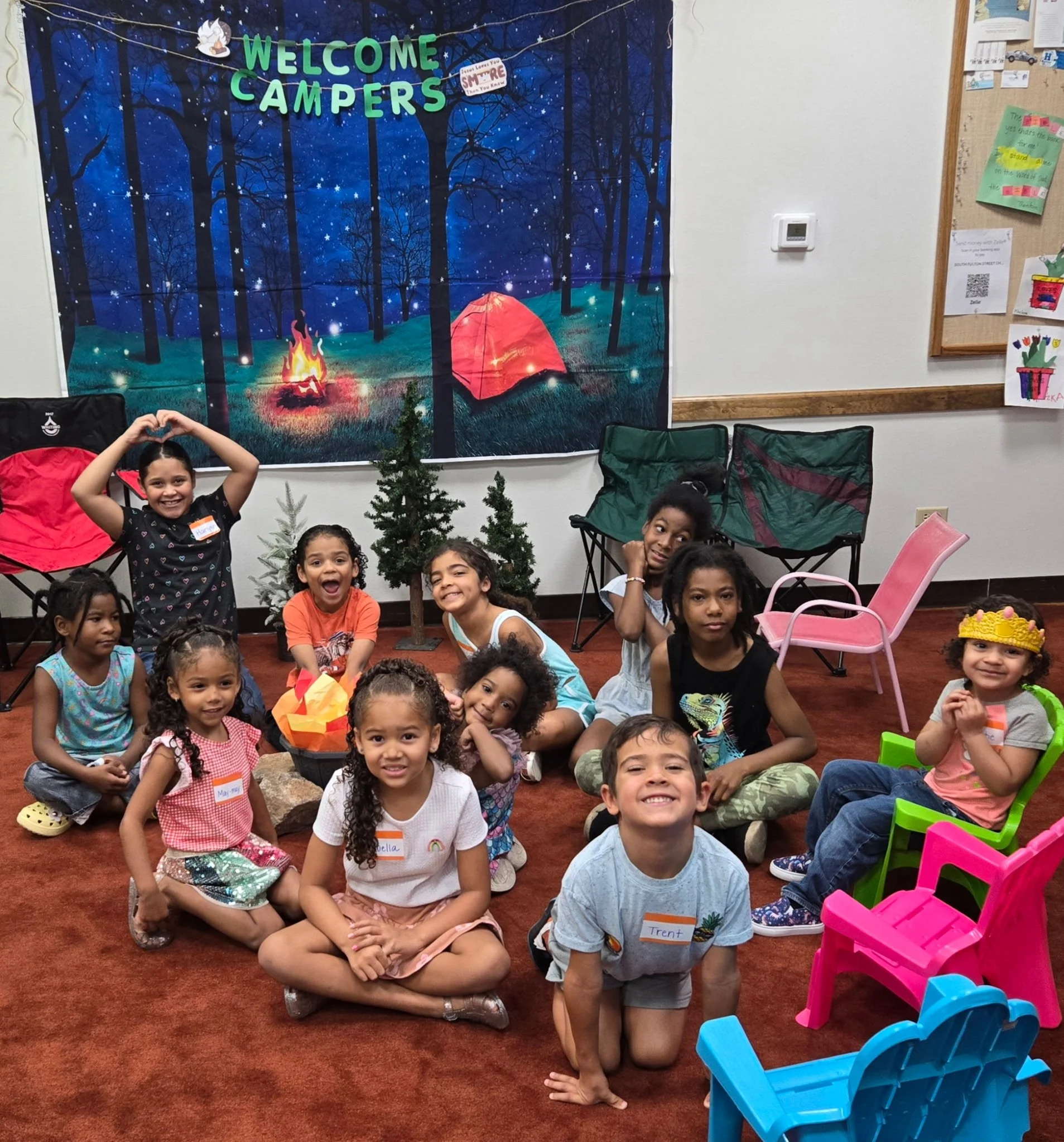 A group of children and a teenager sitting and kneeling on a red-carpeted floor in a classroom, smiling at the camera. Behind them is a large outdoor-themed bulletin board with a forest scene, tents, and a campfire, and a few small potted trees.