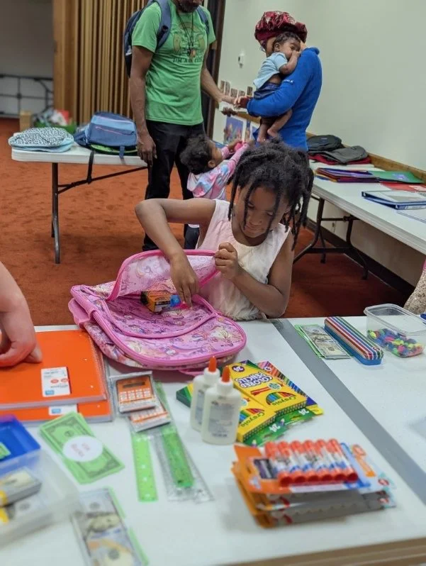A young girl with dreadlocks opens her pink backpack at a table filled with school supplies, while two women and a man stand in the background in a room with tables and colorful folders.