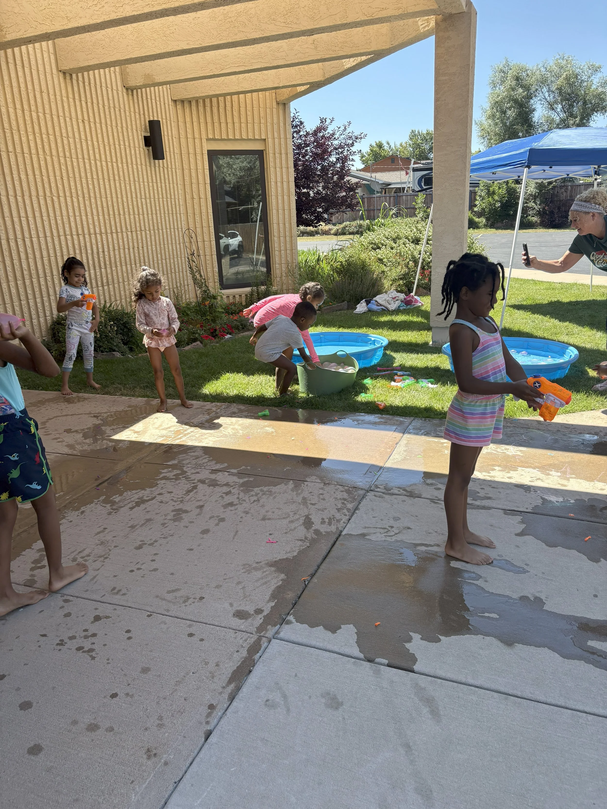 Children playing with water guns and splashing in a backyard on a sunny day, with inflatable pools and a woman taking a photo.