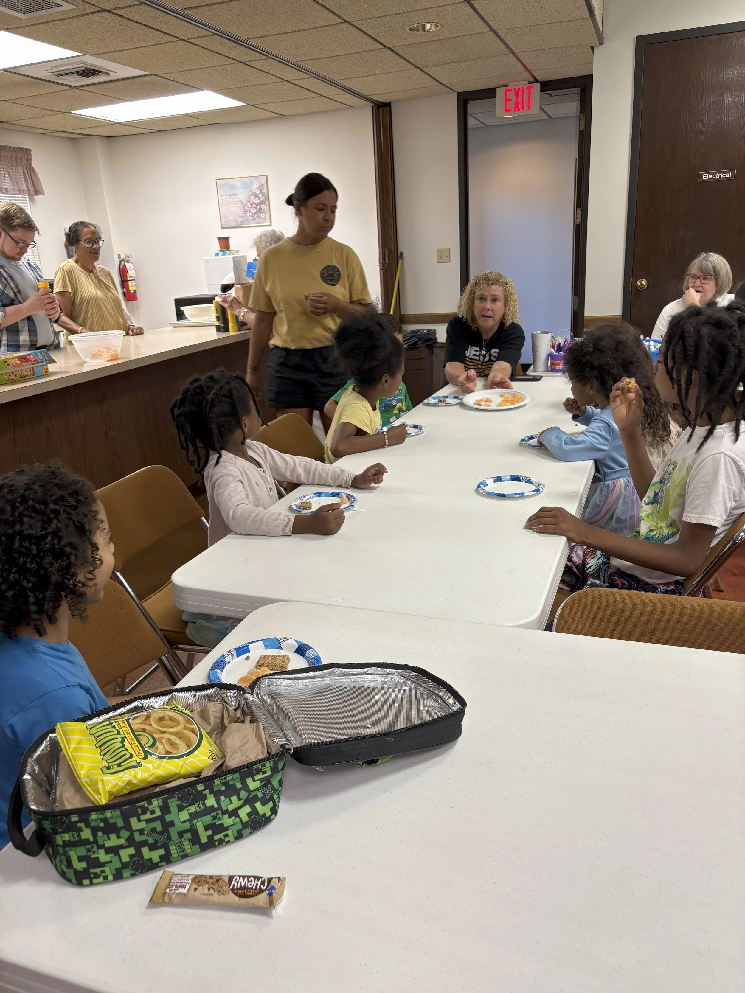 A group of children sitting around a long dining table with plates of food, listening to a woman speaking, while other adults look on in the background in a room with beige walls and wooden accents.