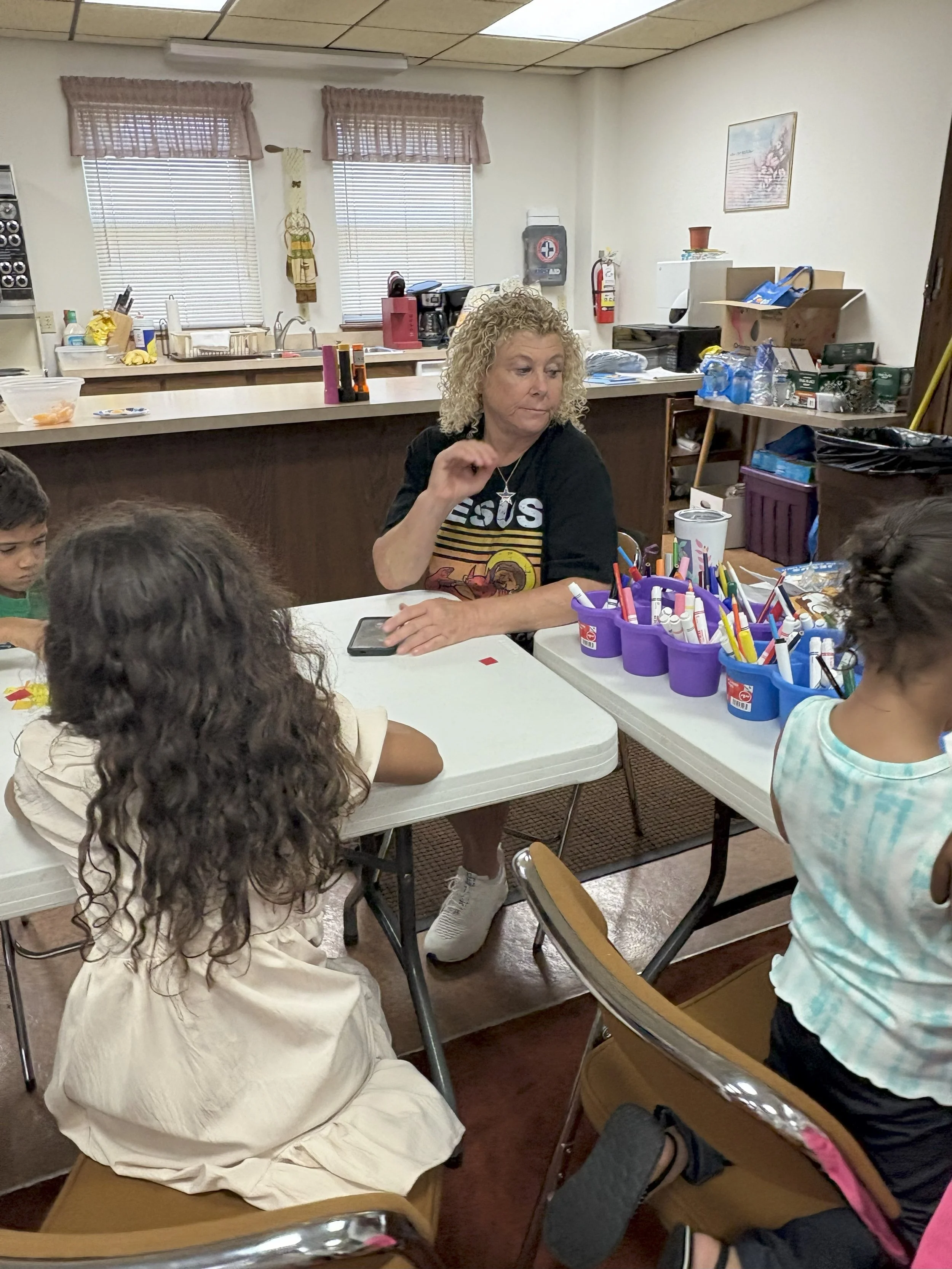 Woman with curly blonde hair sitting at a table with children, engaging in a craft activity with markers, surrounded by supplies in purple and blue cups in a classroom or community room.