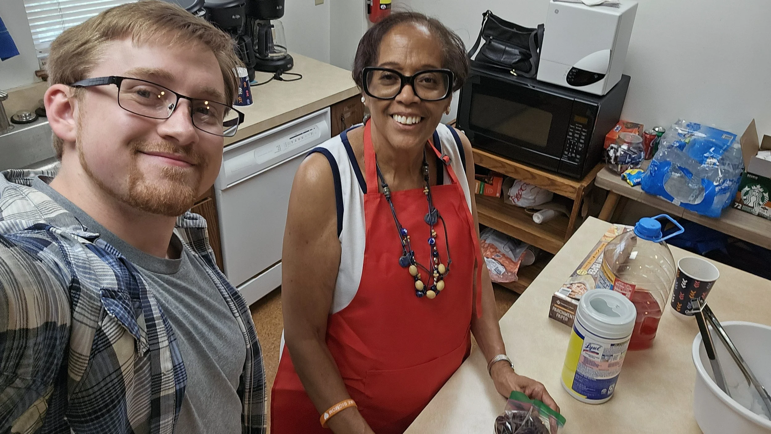 A young man and an older woman smiling in a kitchen, with the woman wearing a red apron, large glasses, and a colorful necklace, and the young man wearing glasses and a plaid shirt. Cooking and kitchen items are visible on the counter and shelves.