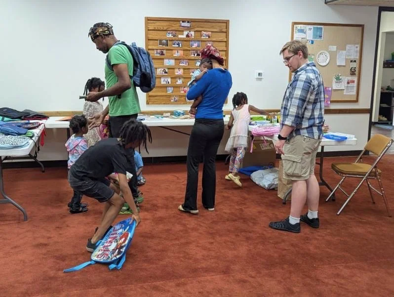 People, including children, standing and sitting in a room with tables displaying clothes and items, a bulletin board, and a chair.