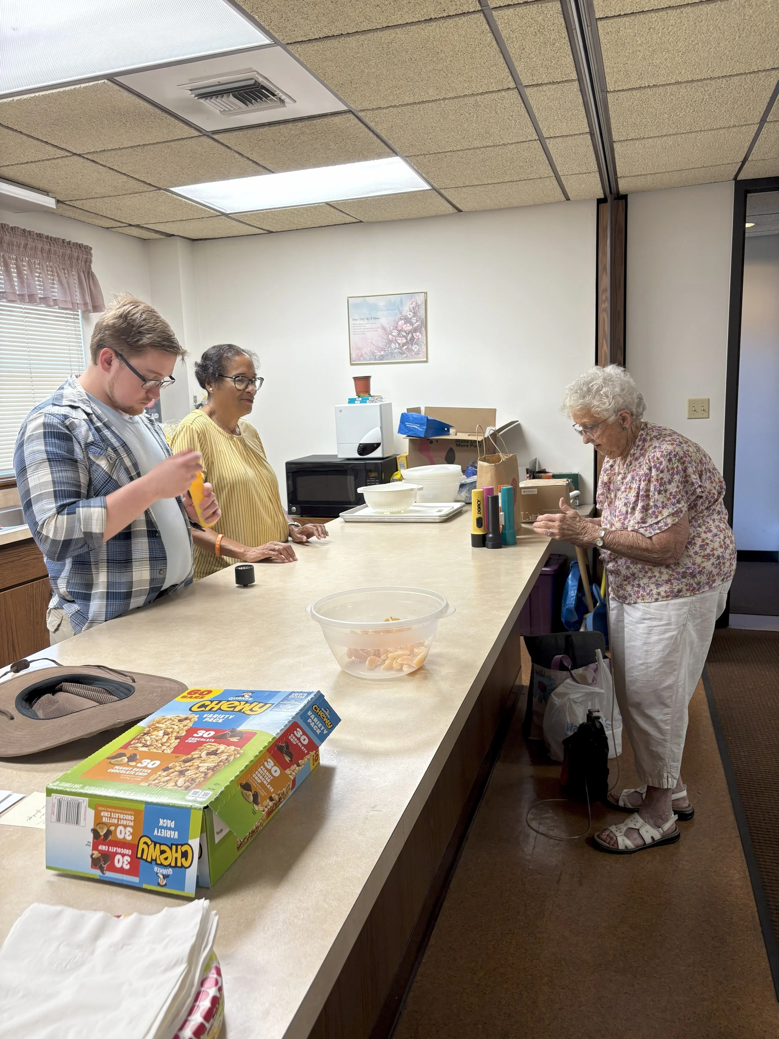 Three people standing at a kitchen counter with food and supplies, an elderly woman on the right and two younger individuals on the left, in a kitchen with white walls and a brown ceiling.