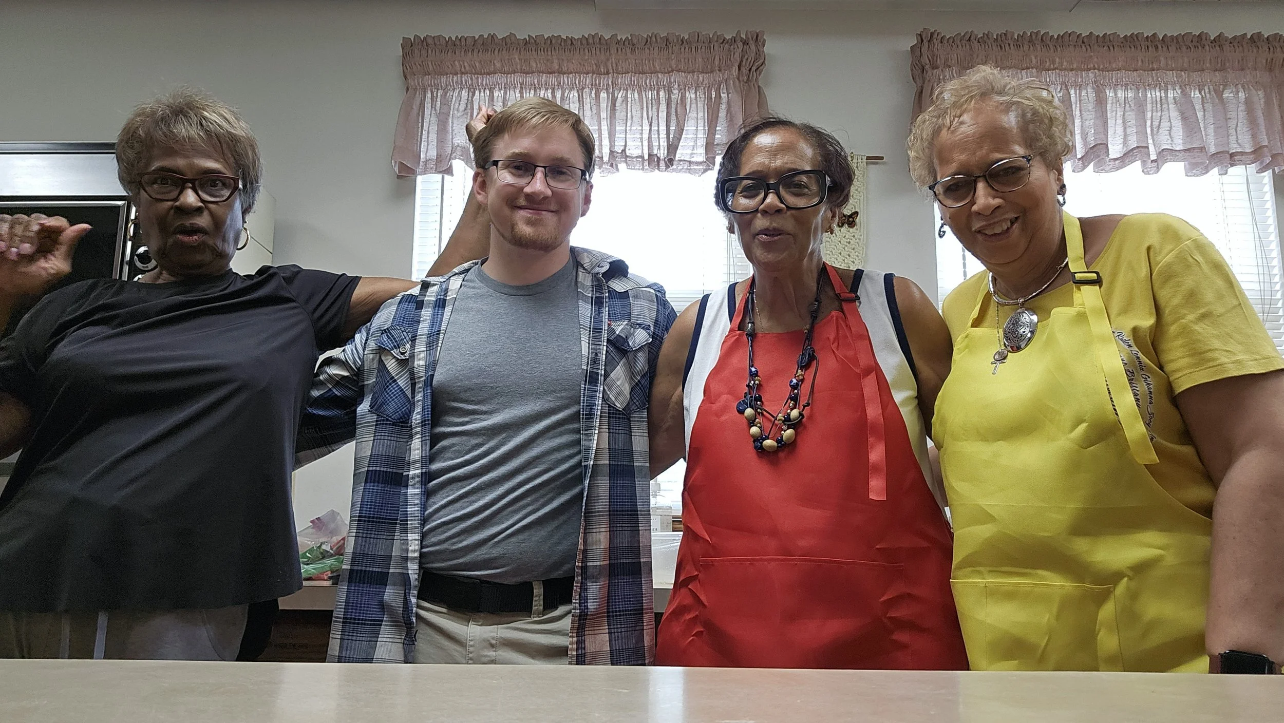 Four women and one young man smiling in a kitchen, standing close together. The women are wearing glasses and colorful aprons, and the young man has glasses and a plaid shirt. They appear to be participating in a cooking or social event.