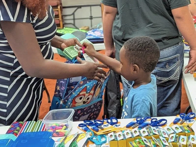 A young boy receiving money from an adult woman at a table with school supplies, including scissors, glue, and notebooks, while another adult stands nearby.