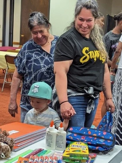 A woman and a young boy holding hands, looking at a table with school supplies and toiletries, in a room with other people.