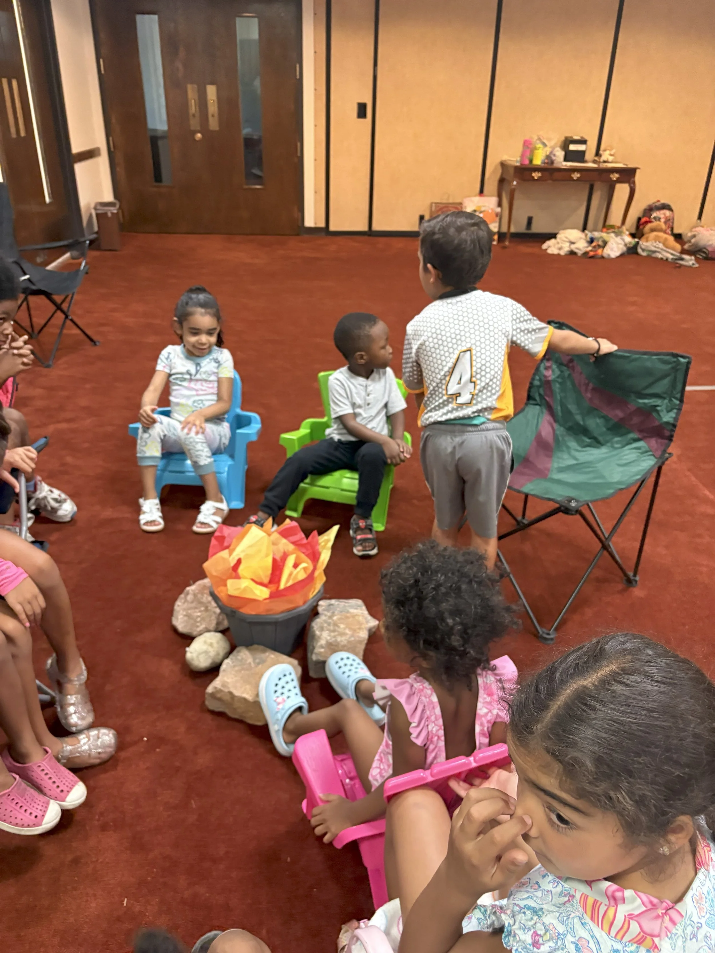 Children sitting and standing in a circle indoors, with some children in chairs and a boy standing in front, possibly giving a presentation or speech.